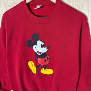 May include: A red sweatshirt with a Mickey Mouse graphic. The cartoon character is in black, white, red, and yellow. The sweatshirt has a crew neck and long sleeves. The garment is suspended on a wooden hanger.