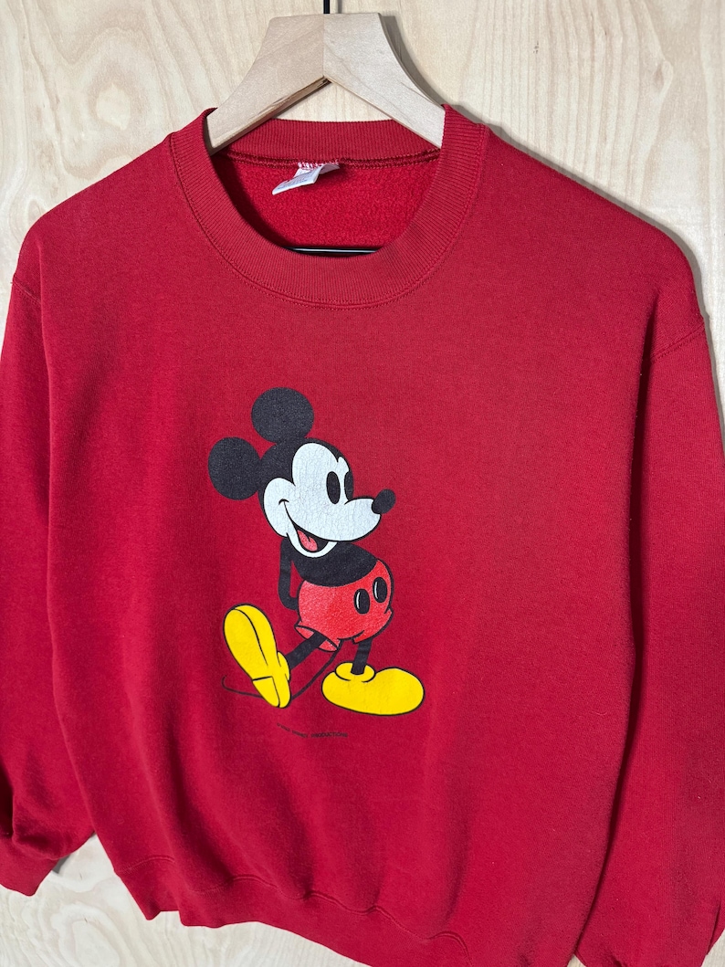 May include: A red sweatshirt with a Mickey Mouse graphic. The cartoon character is in black, white, red, and yellow. The sweatshirt has a crew neck and long sleeves. The garment is suspended on a wooden hanger.