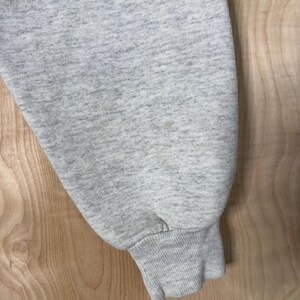 K&ouml;nnte beinhalten: Nahaufnahme eines hellgrauen Sweatshirt-&Auml;rmels. Der &Auml;rmel besteht aus einem weichen, strukturierten Stoff und hat ein geripptes B&uuml;ndchen. Der Hintergrund ist eine hellfarbige Holzoberfl&auml;che.