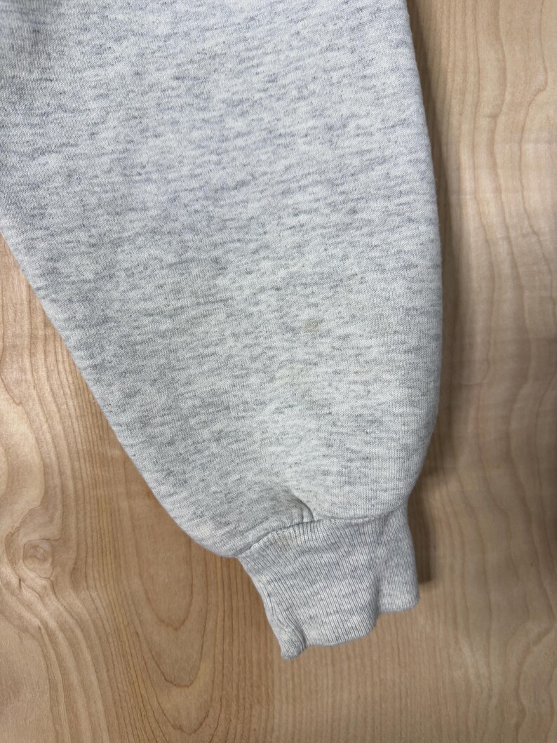 K&ouml;nnte beinhalten: Nahaufnahme eines hellgrauen Sweatshirt-&Auml;rmels. Der &Auml;rmel besteht aus einem weichen, strukturierten Stoff und hat ein geripptes B&uuml;ndchen. Der Hintergrund ist eine hellfarbige Holzoberfl&auml;che.