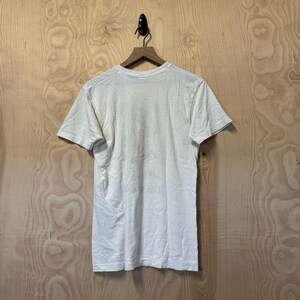 Peut inclure: Un t-shirt cr&egrave;me &agrave; manches courtes est suspendu &agrave; un cintre en bois sur un fond clair &agrave; grain de bois. Le t-shirt est l&eacute;g&egrave;rement froiss&eacute;, sugg&eacute;rant qu'il est fait d'une mati&egrave;re souple. Le dos du t-shirt est visible.