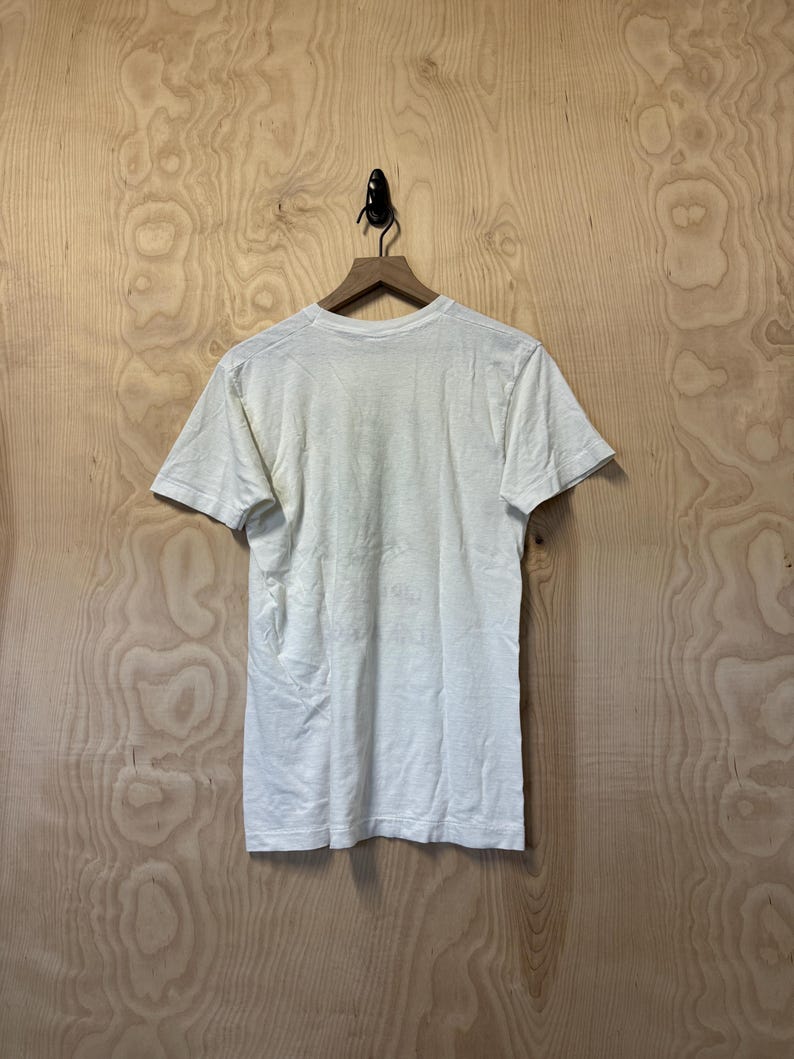 Peut inclure: Un t-shirt cr&egrave;me &agrave; manches courtes est suspendu &agrave; un cintre en bois sur un fond clair &agrave; grain de bois. Le t-shirt est l&eacute;g&egrave;rement froiss&eacute;, sugg&eacute;rant qu'il est fait d'une mati&egrave;re souple. Le dos du t-shirt est visible.