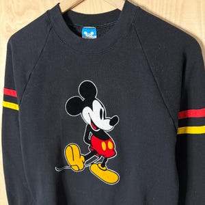 May include: Black sweatshirt featuring a Mickey Mouse graphic. The cartoon character is depicted in his classic pose, with red shorts, yellow shoes, and white gloves. The sleeves have red and yellow stripes. The sweatshirt is hanging on a wooden hanger.
