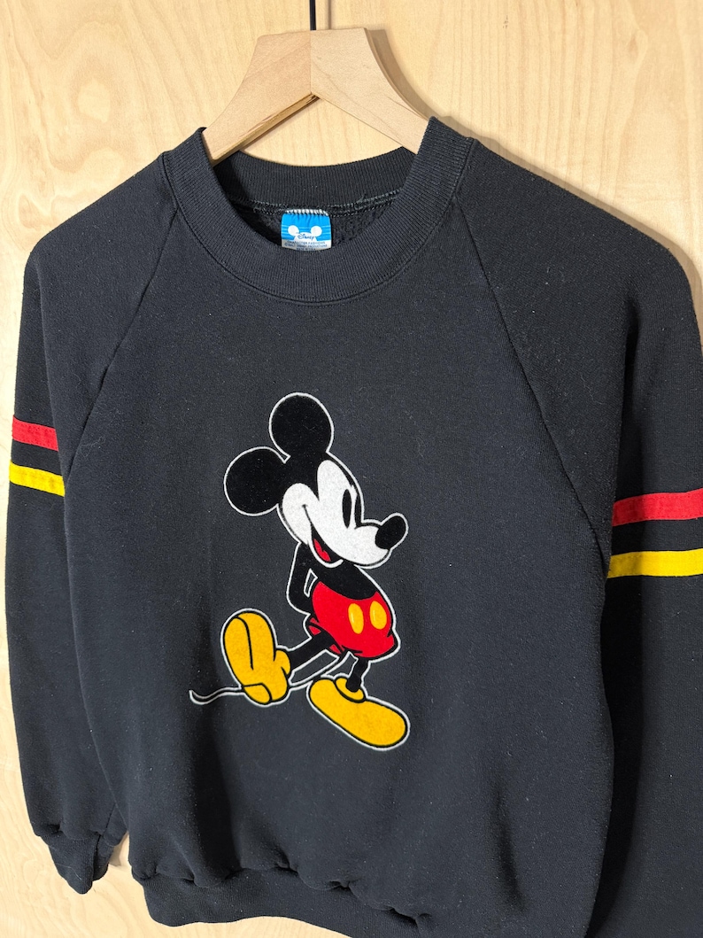 May include: Black sweatshirt featuring a Mickey Mouse graphic. The cartoon character is depicted in his classic pose, with red shorts, yellow shoes, and white gloves. The sleeves have red and yellow stripes. The sweatshirt is hanging on a wooden hanger.
