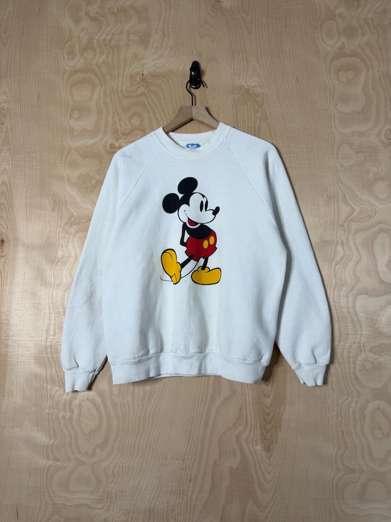 May include: White sweatshirt featuring a large graphic of Mickey Mouse. The cartoon character is depicted in his classic pose, with black ears, a red and yellow outfit, and yellow shoes. The sweatshirt has long sleeves and a crew neck.