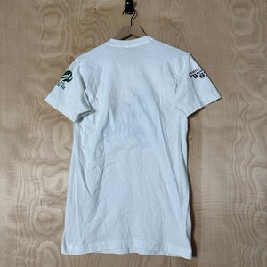 May include: White t-shirt with short sleeves, hanging on a wooden hanger. The shirt has green and white text and a logo on the left sleeve and the words "Central Wyoming" on the right sleeve. The shirt is made of a soft, lightweight material.