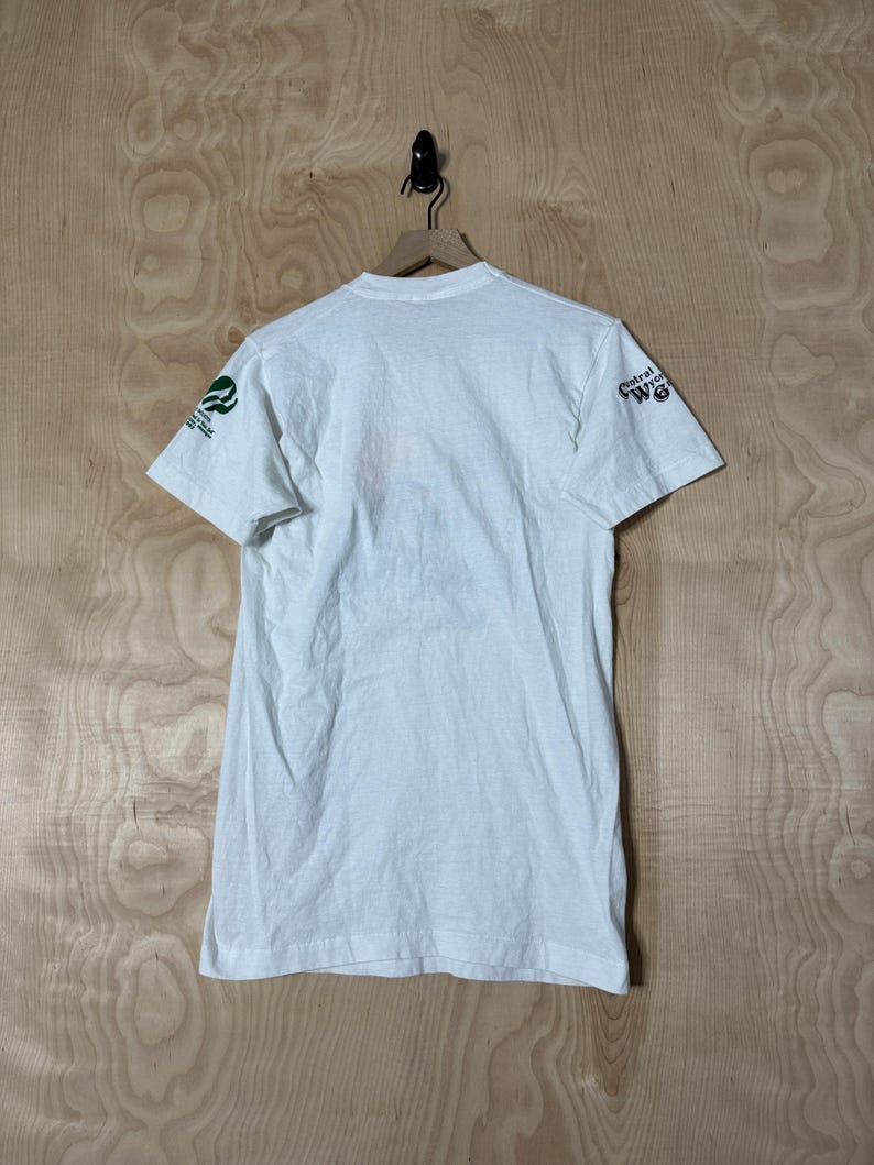 May include: White t-shirt with short sleeves, hanging on a wooden hanger. The shirt has green and white text and a logo on the left sleeve and the words "Central Wyoming" on the right sleeve. The shirt is made of a soft, lightweight material.