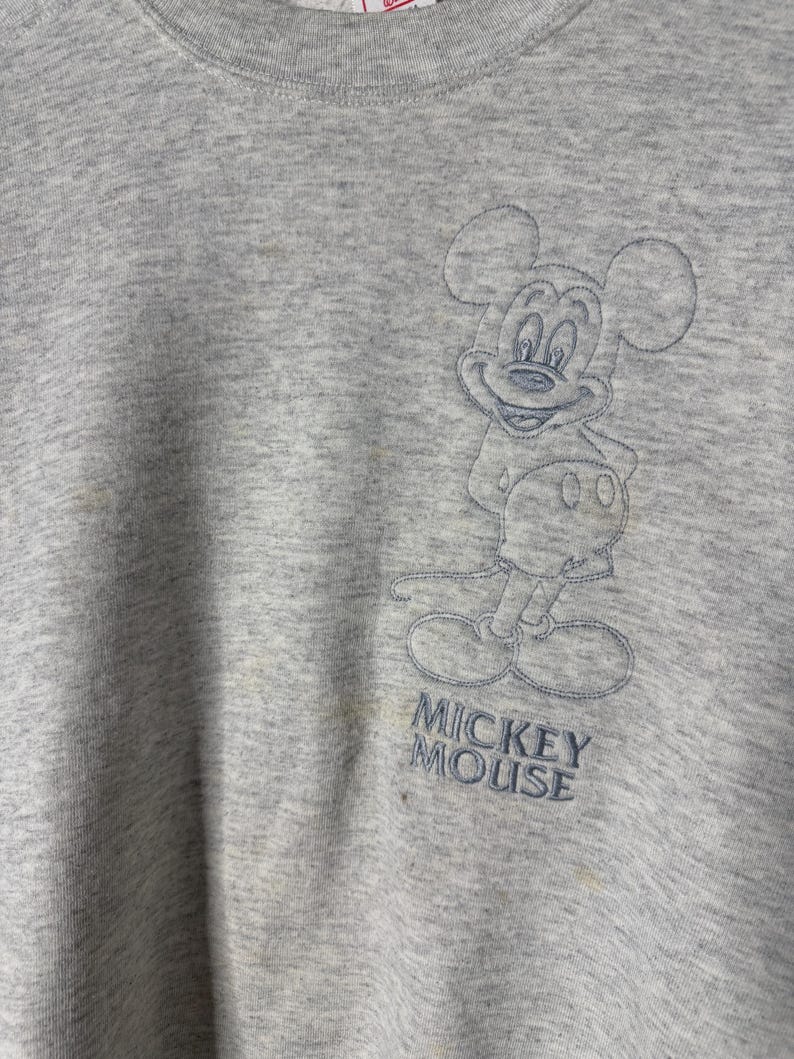 K&ouml;nnte beinhalten: Hellgraues Sweatshirt mit einem gestickten Bild von Mickey Mouse. Der Text "Mickey Mouse" ist unter der Figur gestickt. Das Sweatshirt hat einen Rundhalsausschnitt und lange &Auml;rmel.