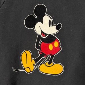 May include: Black sweatshirt featuring a classic Mickey Mouse graphic. The cartoon character is depicted in his iconic pose, with a black head and ears, white face, red shorts, yellow shoes, and white gloves. The design is outlined in white.