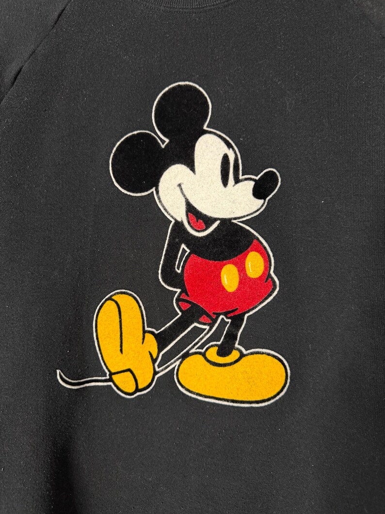 May include: Black sweatshirt featuring a classic Mickey Mouse graphic. The cartoon character is depicted in his iconic pose, with a black head and ears, white face, red shorts, yellow shoes, and white gloves. The design is outlined in white.