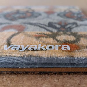 May include: A rectangular mousepad with a floral pattern in shades of gray, orange, and beige. The word "vayakora" is printed in white on the lower portion of the mousepad. The edges are a dark gray.