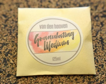 Könnte beinhalten: Kleines, goldfarbenes Päckchen mit rundem Etikett. Das Etikett trägt die Aufschrift "van den hooven Granulating Medium" in schwarzer Schrift über einem Aquarelldesign. Der Boden des Etiketts zeigt "125ml".
