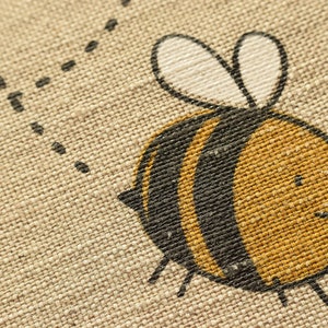 Puede incluir: Una abeja amarilla y negra con alas blancas vuela sobre un fondo de lino beige. La abeja tiene una cara sonriente y vuela en un patrón de línea punteada.