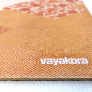 May include: Close-up of a rectangular, patterned surface with a geometric design in shades of orange and tan. The word "vayakora" is printed in white. The surface has a textured appearance.