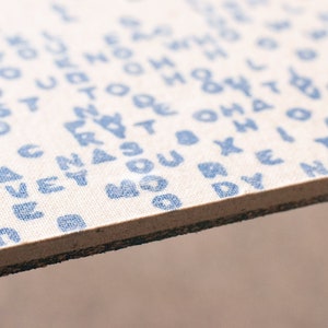 Puede incluir: Primer plano de un trozo rectangular de tela con texto azul impreso. El texto parece ser una variedad aleatoria de palabras y frases. La tela es de color beige claro y tiene un borde marrón oscuro.