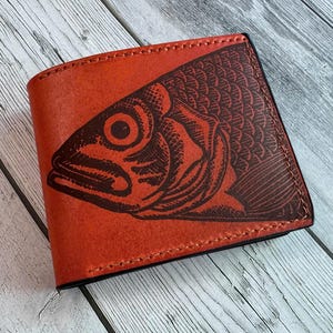 May include: A bi-fold wallet made of orange leather featuring a dark brown embossed graphic of a fish head on the front cover. The wallet has visible stitching along the edges and a smooth, matte finish on the exterior surface.