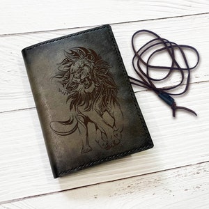 May include: A brown leather journal with a lion design embossed on the front. The journal has a brown leather strap with a metal clasp.