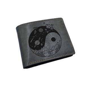 Puede incluir: Una cartera gris de dos pliegues con un diseño circular grabado en láser negro. El gráfico muestra un símbolo de yin-yang compuesto por una escena espacial con planetas y asteroides en un lado y un sol sobre un paisaje montañoso en el otro.