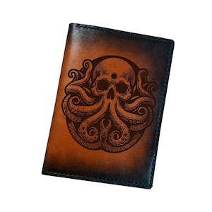 May include: A rectangular leather passport holder featuring a dark brown and black gradient finish. The front surface displays a detailed, dark-toned laser-engraved illustration of a human skull integrated with octopus tentacles in a circular design.
