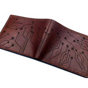 May include: A brown leather bifold wallet featuring a black laser-engraved circuit board pattern across the exterior surface. The wallet has visible stitching along the edges and a smooth, matte finish on the leather material.