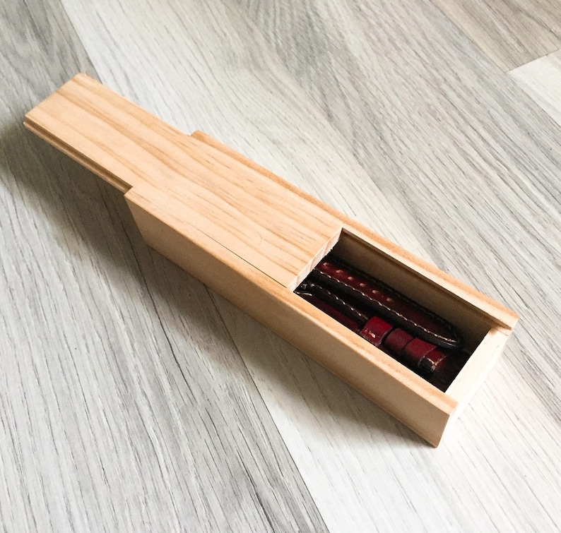 Puede incluir: Una caja de madera con una tapa deslizante, que revela dos correas de reloj de cuero marr&oacute;n en el interior.