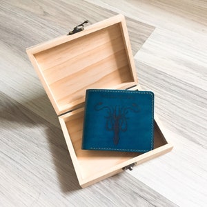 Puede incluir: Una cartera de cuero azul verdoso con un dise&ntilde;o negro en relieve de un drag&oacute;n de tres cabezas. La cartera est&aacute; en una caja de madera.