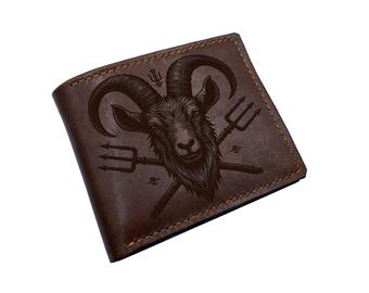May include: A brown leather bifold wallet featuring a dark laser-engraved illustration of a goat head centered between two crossed tridents. The wallet has visible stitching along the edges and a smooth, matte finish on the front surface.