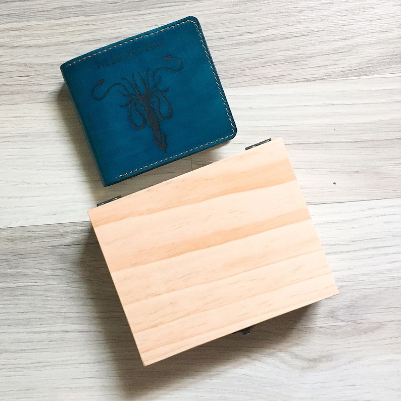 Puede incluir: Una cartera de cuero azul verdoso con un gr&aacute;fico en blanco y negro de un calamar y el texto "The Great Kraken". La cartera est&aacute; encima de una caja de madera marr&oacute;n claro.