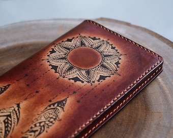 May include: A brown leather wallet with a detailed mandala design and feather motifs. The wallet is hand-stitched with a contrasting thread and has a rich, textured appearance. The design is etched in black, creating a striking contrast.