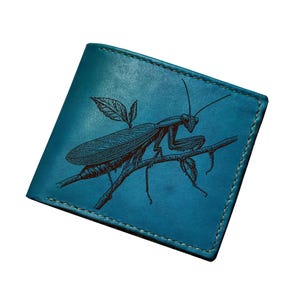 May include: A teal leather bifold wallet featuring a dark brown engraved illustration of a praying mantis perched on a small branch with leaves. The wallet has visible stitching along the edges and a smooth, matte finish on the exterior surface.