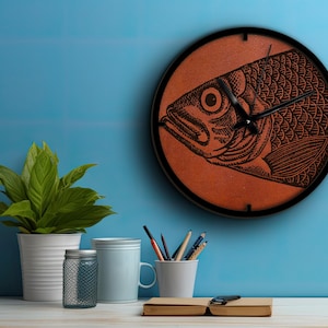 May include: A round wall clock with a black frame and a brown background. The clock face features a black and white illustration of a fish.