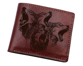 May include: A brown leather bifold wallet featuring a dark, engraved graphic of three howling wolves with open mouths and visible teeth. The wallet is constructed with visible tan stitching along the top and side edges, showcasing a smooth finish.