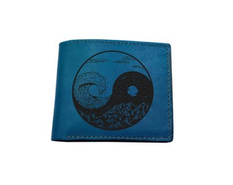 Op de afbeelding: Een blauwe portemonnee met een zwarte cirkelvormige afbeelding op de voorzijde. Het ontwerp toont een yin-yang verdeling met een brekende oceaangolf aan de ene kant en een nachtelijke hemel met een maan en bergketen aan de andere kant.