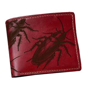 Könnte beinhalten: Ein tiefrotes Bifold-Portemonnaie mit einem dunkelbraunen, eingravierten Muster aus mehreren Kakerlaken auf der Vorderseite. Das Portemonnaie ist mit sichtbaren hellen Nähten an den Kanten versehen und hat eine glatte, polierte Oberfläche.