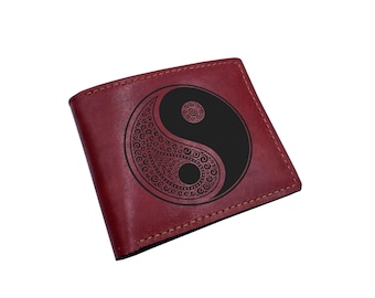 Op de afbeelding: Een donkerrode bifold portemonnee met een zwart yin en yang symbool op de voorzijde. Het yin en yang ontwerp is versierd met ingewikkelde wervelende patronen en kleine stippen. De portemonnee heeft zichtbare stiksels langs de randen.