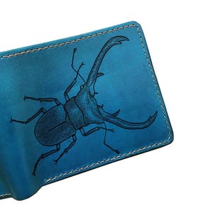 May include: A bright blue leather bifold wallet featuring a detailed black line art illustration of a stag beetle on the front cover. The wallet is constructed with visible light-coloured stitching along the edges and has a smooth texture.