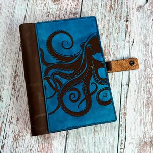 Può includere: Un diario in pelle blu con una rilegatura in pelle marrone e un design di polpo marrone in rilievo sulla copertina. Il diario ha un cinturino in pelle marrone con un bottone metallico.