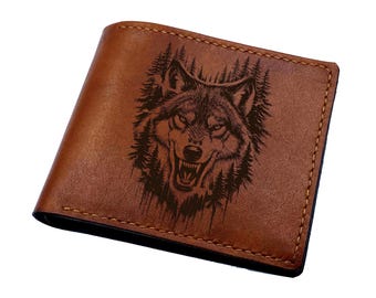 May include: A brown leather bifold wallet featuring a dark, laser-engraved illustration of a snarling wolf head surrounded by a dense forest of pine trees. The wallet has visible stitching along the edges and a smooth, matte-textured surface.