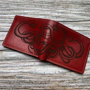 Peut inclure: Un portefeuille bifold rouge foncé orné d'un motif gravé au laser représentant un crâne entrelacé de plusieurs serpents. Le portefeuille présente des coutures visibles sur les bords et une surface en cuir lisse et polie sur l'extérieur.