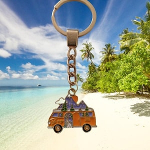 May include: A gold-colored metal keychain featuring a cartoon camper van with a mountain scene and a blue sky. The van is orange with a white door and windows. The mountains are purple and blue with green trees. The keychain is attached to a silver chain and a silver ring.