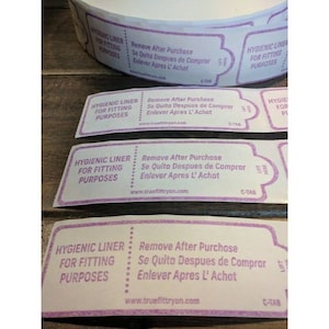 May include: Four pink and white labels with the text "Hygienic Liner for Fitting Purposes" and "Remove After Purchase Se Quita Despues de Comprar Enlever Apres L' Achat" and the website "www.truefittryon.com". The labels are on a roll and have a C-Tab.