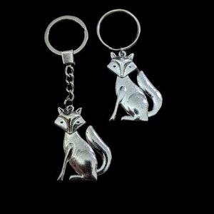May include: Two silver fox keychains. One keychain has a chain, the other has a simple ring. Both keychains feature a detailed fox design with a sitting pose and a curved tail. The keychains are set against a black background.