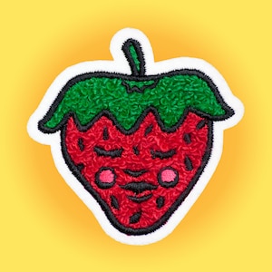 Può includere: Un toppa ricamata a forma di fragola rossa e verde con un viso sorridente. La fragola ha una texture pelosa e contorni neri.