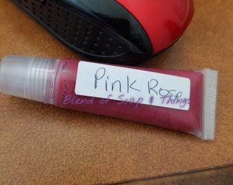 May include: A clear tube with a pink lip gloss inside. The tube has a white label with the text "Pink Rose" and "A Blend of Soap & Things" written on it.