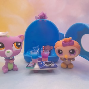 May include: A scene featuring Littlest Pet Shop figures. A purple cat figure with a white bow sits next to a table with drinks and a phone. An orange owl figure stands nearby. A blue carrying case is in the background.