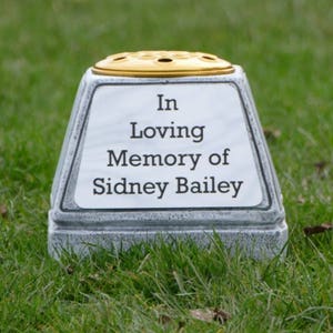 May include: A memorial vase with a white panel inscribed with "In Loving Memory of Sidney Bailey." The vase has a grey stone-like base and a gold-coloured top with holes. It is set on green grass.