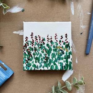 May include: A small square canvas painting featuring a stylized landscape. The artwork depicts green brushstrokes representing foliage, with red berry-like accents. The background is a neutral off-white, and the painting is set against a brown surface.