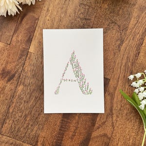 May include: A white card features the letter "A" artfully designed with green leaves and small pink flowers. The card is set on a wooden surface, with white flowers and greenery in the background.