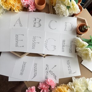May include: White cards with the letters A through L, each decorated with a floral design. The cards are surrounded by yellow and pink flowers, small terracotta pots, and a wooden tray.