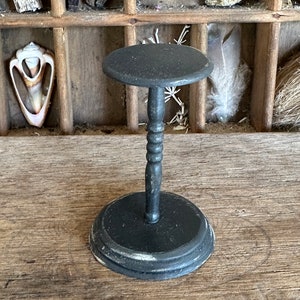 Puede incluir: Un soporte para sombreros de madera negro con una parte superior redonda y una base de pedestal torneada. El soporte está desgastado con un acabado de pintura blanca.